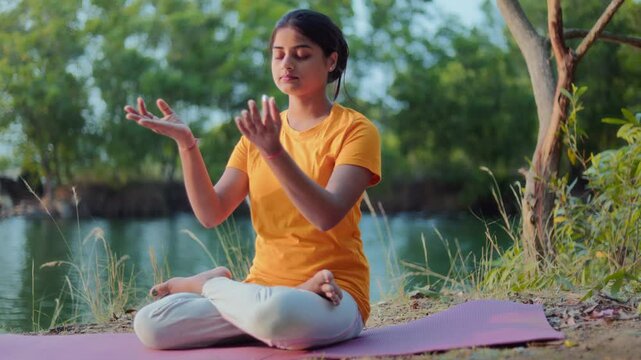 Young Indian woman sitting in padmasana or lotus position with gyana mudra and changing it to prayer pose by a calm lake.