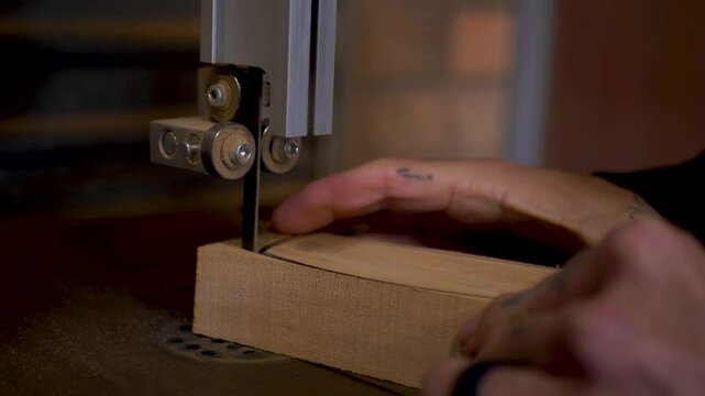 Slow motion close-up of man with hand tattoos carefully cutting wooden climbing hold blocks with electric bandsaw machinery blade tool DIY home garage woodwork shed in Australia carpentry industry