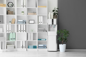 Modern water cooler with paper cups and shelf unit in office