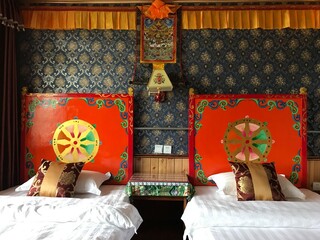 Tibetan - style hotel room with traditional decor, double beds, ethnic headboards featuring Dharma wheels, traditional Tangka display on classic Tibetan - pattern wallpaper.