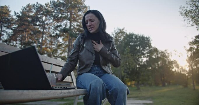 A woman sitting on a park bench with her laptop suddenly feels unwell. She experiences stomach pain and chest discomfort, struggling with an unexpected illness while trying to work remotely in nature.