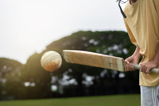 Young boy wearing a yellow shirt and helmet plays cricket on a grassy field, focusing intensely as he hits the ball with a wooden bat. Outdoor sports and youth energy captured in motion.