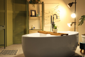 Interior of bathroom with bathtub, green folding screen and glowing lamps in evening