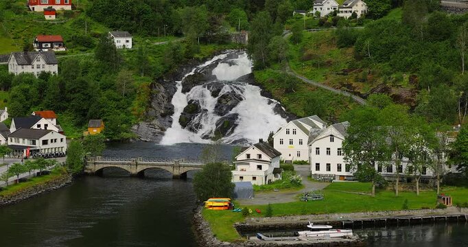 View on Hellesylt, a small village in the Sunnylvsfjord, Norway. Captured from a large cruise ship, the scene features a lush green landscape, a waterfall, and serene fjord waters.