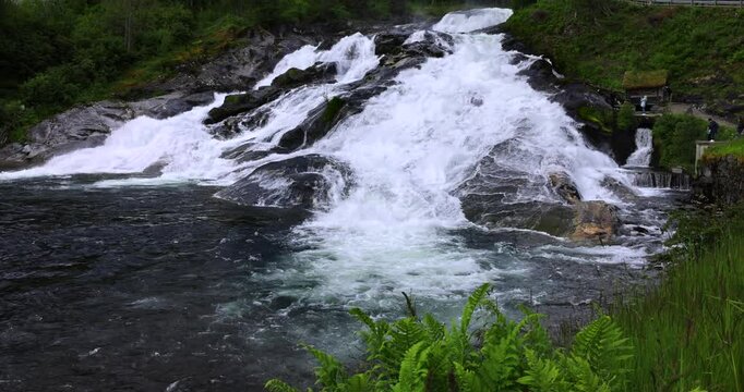 Powerful waterfall near Hellesylt, Norway, cascading over dark rocks into a crystal-clear river, surrounded by lush greenery, captured during a fjord cruise excursion.