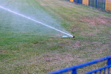Automatic sprinkler irrigating sports field with dry grass patches, highlighting the need for watering to prevent drought damage and showing impact of climate change on green spaces.