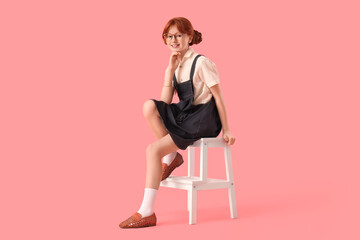 Cute ginger girl in stylish dress and eyeglasses sitting on chair against pink background