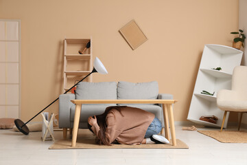 Scared young woman hiding under table and covering head in living room during earthquake