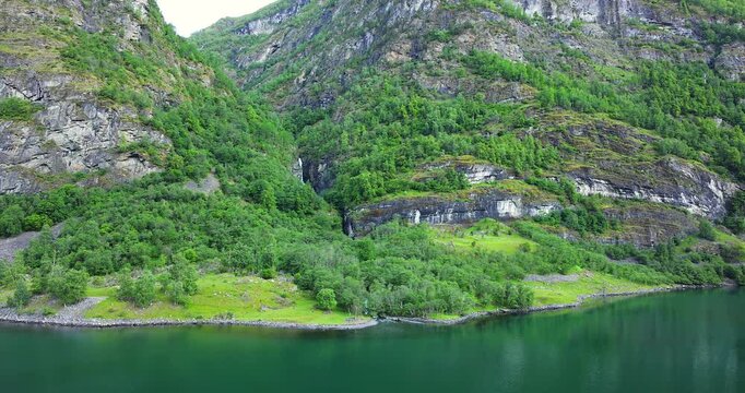 Majestic green cliffs and deep blue fjord waters under cloudy skies, filmed from a cruise ship sailing through the breathtaking landscapes of Norway fjords.