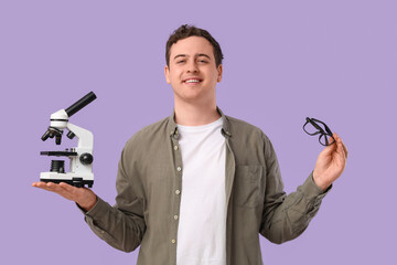 Handsome young man with microscope and eyeglasses on lilac background