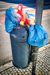 Overflowing waste bin on a city street with plastic bags and litter during daylight hours