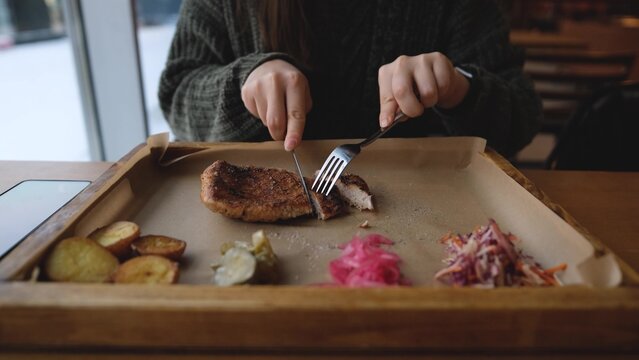 Pretty brunette enjoying tasty beef steak in restaurant in winter. Woman in warm cozy sweater eating meat near window. Snow outside