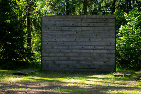 A tall wooden climbing wall with handholds stands in a sunlit forest, likely part of an outdoor obstacle or training course.
- Powered by Adobe
