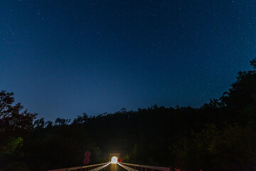 A starry night sky above a dark road leading into a tunnel, framed by dense trees and a shooting...