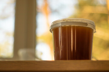 A plastic container filled with honey sits on a wooden surface, with warm natural light and soft...