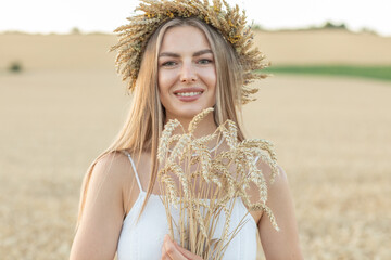 Portrait of a beautiful young woman in white dress and wreath of wheat ears in the field.