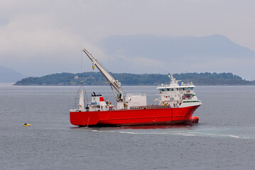 A large red supply vessel moves slowly through calm waters with an island in the background under...