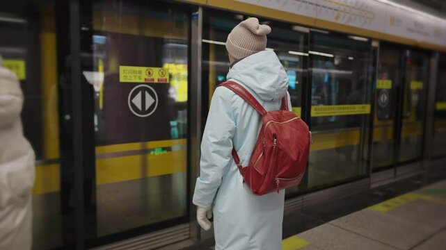 A young woman in a blue raincoat and red backpack waits for a commuter train in a modern Asian subway station, likely in China.