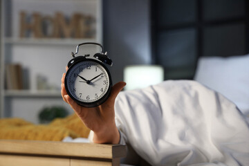 Young woman under blanket holding retro alarm clock in bedroom, closeup