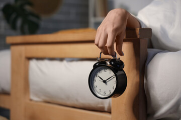 Young woman under blanket holding retro alarm clock in bedroom, closeup