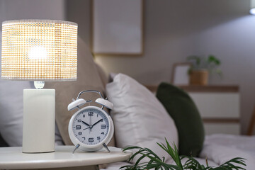 Alarm clock and glowing lamp on bedside table in bedroom, closeup