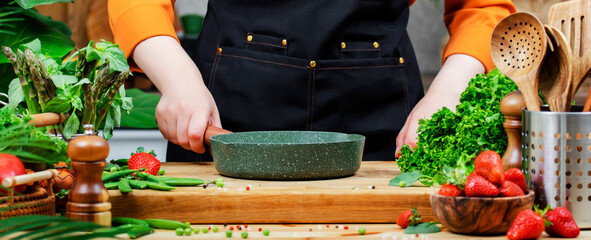 woman holds green frying pan in hands prepares healthy vegan meals in kitchen, surrounded by fresh...
