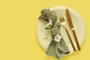 Beautiful table setting with plate, golden cutlery and flowers on yellow background