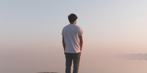 Young man standing on rocky shore, gazing at tranquil water during sunset, surrounded by soft pastel colors, reflecting calmness and introspection in nature's beauty