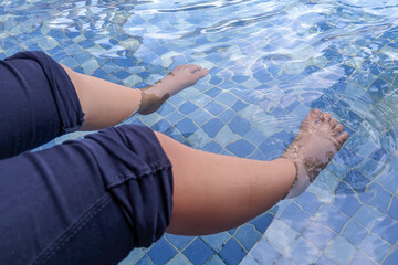 A person's legs and feet are partially submerged in clear, cool pool water with blue and white tiles, capturing a moment of relaxation.