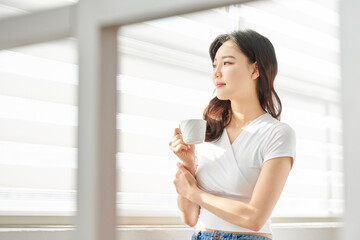 Young Asian Female Standing by Window Holding Coffee Cup in Bright Morning Light
