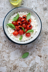 Bowl of cottage cheese caprese with roasted tomatoes and green basil, vertical shot on a brown granite background with space, view from above
