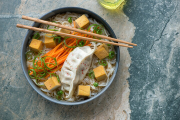 Bowl of rice noodles with smoked tofu and steamed dumpling, horizontal shot on a beige and grey...