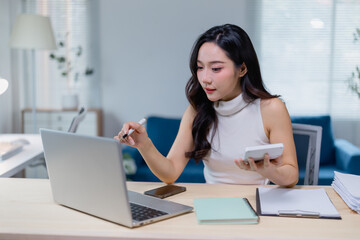 Obraz premium Young professional woman multitasking at her desk, using a laptop and calculator for business tasks. Modern office setting with natural light and organized workspace