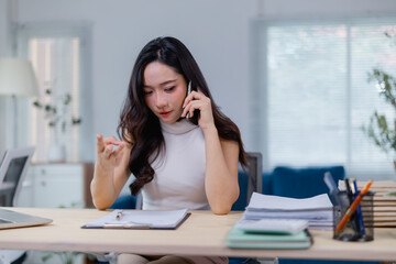 Obraz premium Focused young woman multitasking in a modern office, speaking on the phone while reviewing documents. The workspace is organized, with a laptop, papers, and office supplies neatly arranged