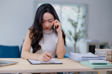 Professional woman managing tasks efficiently by talking on the phone and writing notes at her desk. Office environment with documents and laptop visible in the background