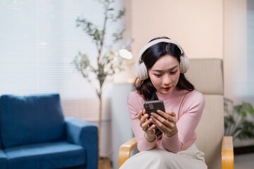 Young woman wearing headphones is focused on her smartphone while sitting indoors. The setting is...