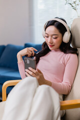 Young woman with headphones relaxes in a cozy chair, engaging with her smartphone. She enjoys the...