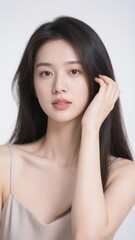 Fototapeta premium A person with long dark hair gently touching their face, wearing a light-colored sleeveless top against a neutral background.