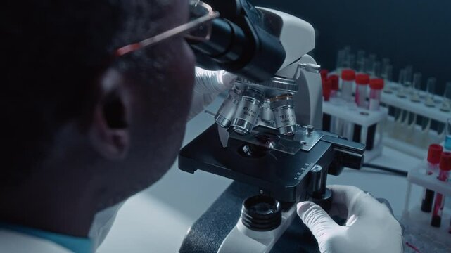Over-shoulder close-up of black male laboratory technician turning revolving nosepiece on electronic microscope, then adjusting stage with blood specimen, while performing bloodwork tests
