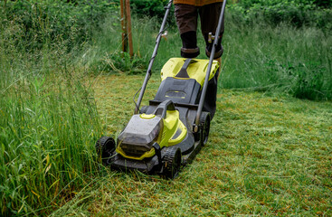 Man mowing overgrown lawn with yellow electric mower near wooden house steps