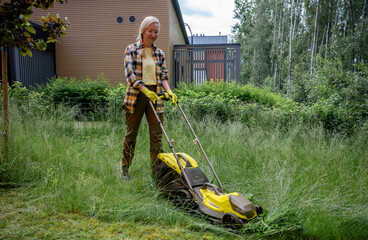 Woman using electric lawn mower in overgrown garden near wooden house