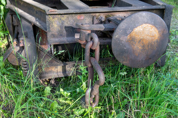 Close-up of rusty coupling chain and buffer on abandoned narrow gauge railway wagon overgrown with grass in summer forest, symbol of decay and forgotten transportation