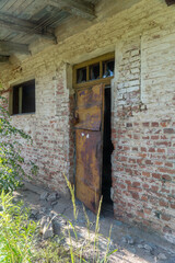 Abandoned brick building with rusted metal door slightly open, broken window panes, and crumbling...