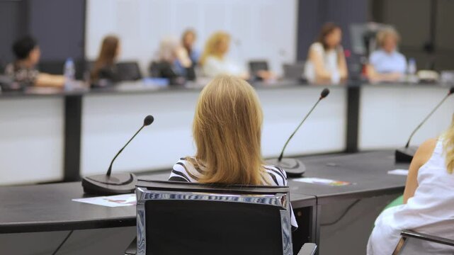 Woman sits in a chair at a conference table. She is wearing a striped shirt and has a microphone in front of her. There are several other people in the room