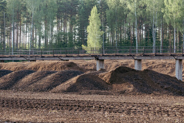 Peat mounds in rural industrial landscape with concrete railway bridge, forest in background,...
