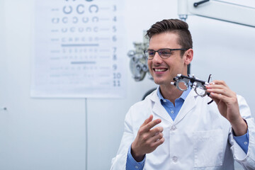 Trial frame sitting on exam table in optometry clinic, with phoropter arm and blurred Snellen chart