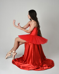 female ballerina dancer model, graceful seated pose on red silk chair, stretching with graceful arms. wearing classical ballet red tutu and pointe shoes costume on white isolated studio background