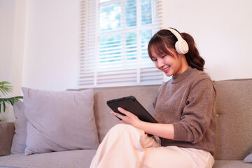 Young Asian adult woman relaxed on couch using tablet and headphones for entertainment digital content consumption daily modern life