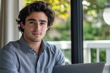 Fototapeta premium Young man with curly hair, wearing a grey shirt, sits near a window with a laptop.