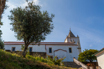 A white portuguese church with a red-tiled roof and a tall steeple stands gracefully amidst greenery